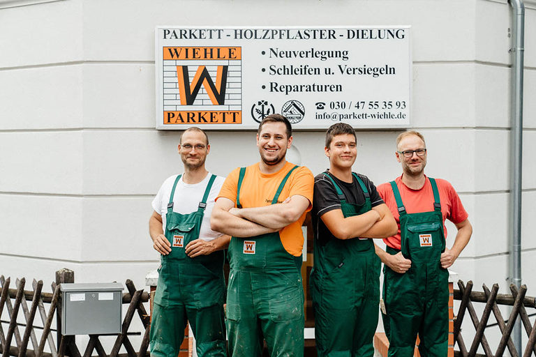 Vier Personen - Parkettlegermeister und Azubis - posieren vor ihrer Parkettwerkstatt.