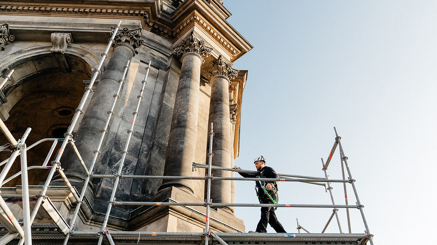 Ein Gerüstbauer läuft auf einem Gerüst entlang mit einer Gerüststange. Das Gerüst befindet sich an einem historischen Gebäude in Berlin. 