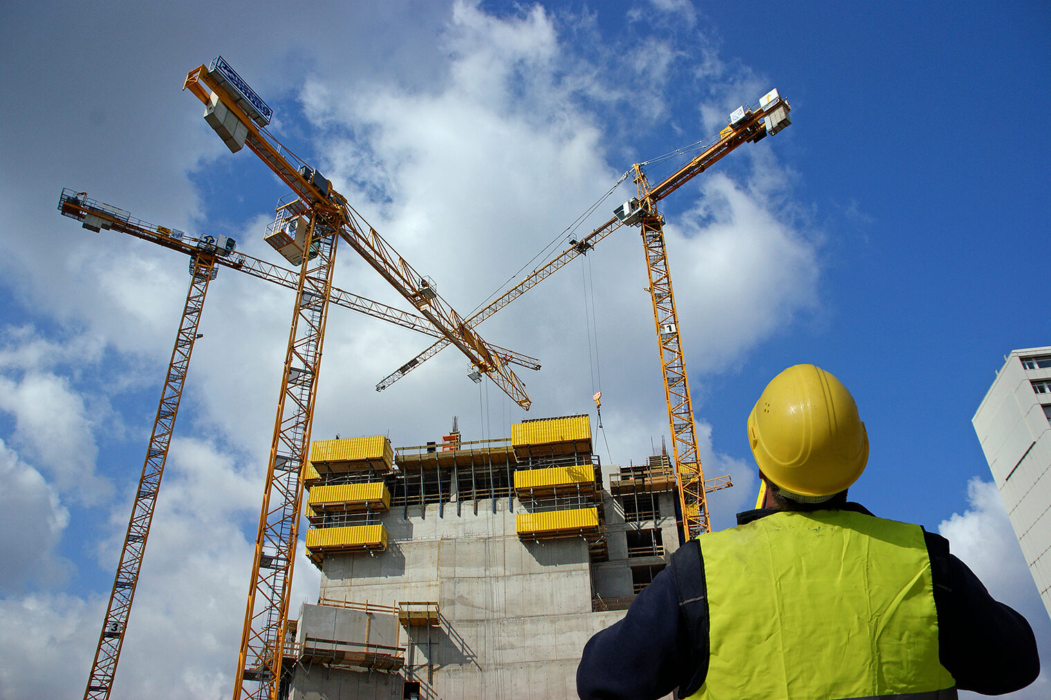Ein Bauarbeiter mit Helm steht vor einem Neubau auf einer riesigen Baustelle und schaut auf drei Kräne.